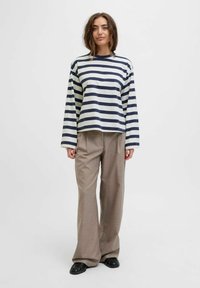 A woman stands confidently, wearing a striped long-sleeve shirt and loose plaid pants, with a neutral background emphasizing her outfit.