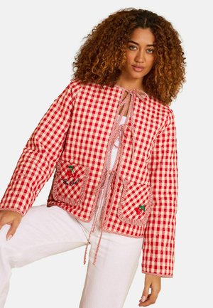 Naisel käharate juustega, kes kannab punase-valge ruudulise mustriga jopet lipsukinnitustega ja tikanditega taskute ning valgete pükste peal.