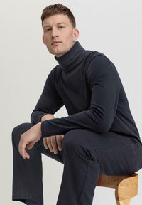 Marineblaue Rollkragenpullover aus weichem Material, mit langen Ärmel und taillierter Passform, kombiniert mit dunklen Hosen, sitzt auf einem Holzstuhl.