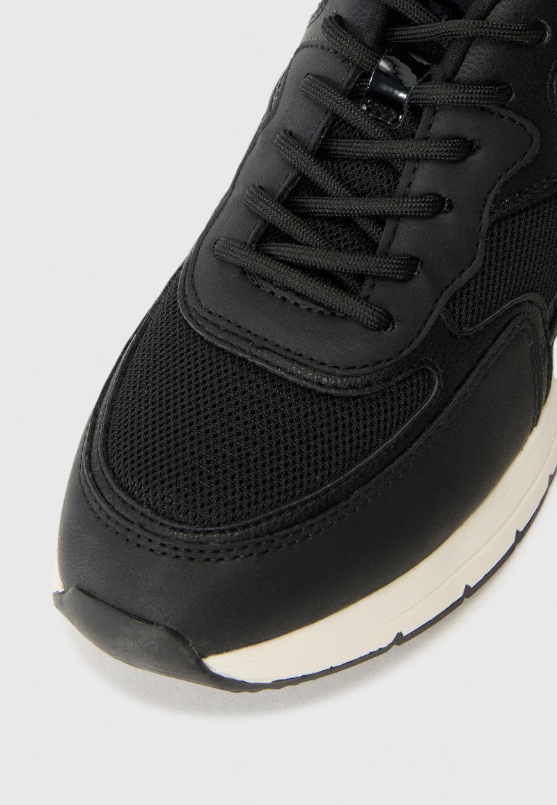 Baskets noir avec une tige en maille texturée, des accents en cuir lisse, un bout rond et une semelle intermédiaire blanche contrastante avec des détails texturés sur la semelle.