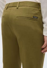 Παντελόνι ελιάς από υφασμένο ύφασμα με υφή, με δύο πίσω τσέπες και μια μικρή ετικέτα. Κομψή γραμμή με θηλιές για ζώνη στη μέση.