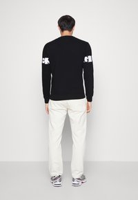 Homme vu de dos, portant un haut à manches longues noir avec du texte blanc sur les manches, un pantalon blanc et des chaussures de course argentées, sur un fond uni.