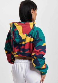 Skrajni hoodie z večbarvnim vzorcem kamuflaže v zelenih, rdečih, rumenih in mornarsko modrih odtenkih. Vključuje kapuco in elastičen rob.