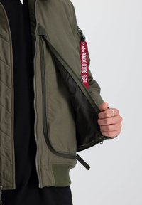 Olivovo zelená bombrová bunda s prešívaným dizajnom, s červenou visačkou "REMOVE BEFORE FLIGHT", so zipsovými vreckami a rebrovanými manžetami.