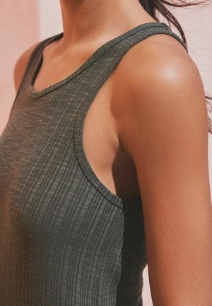 Nærbillede af en person iført en ribbet, mørkegrå, ærmeløs top med fokus på skulder og overarm mod en lys baggrund.