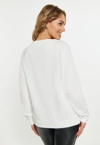 Witte sweatshirt met raglanmouwen, geribbelde halslijn en Zoom. Glad textuur, eenvoudig ontwerp en getailleerde silhouette. Draagt bij zwarte leggings.
