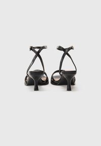 Schwarze Absatzsandalen mit offenem Rücken, ausgestattet mit überkreuzten Riemen und einem strukturierten Zehenbereich. Der Absatz ist kurz und stabil für einen sicheren Halt.