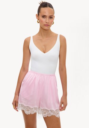 Femme avec les cheveux en chignon porte des boucles d'oreilles créoles dorées, un top blanc sans manches et un short en satin rose clair avec une bordure en dentelle blanche.
