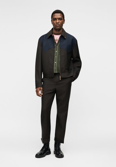 Άνδρας που φοράει μαύρο και ναυτικό μπουφάν με φερμουάρ πάνω από πράσινο πουκάμισο με κουμπιά, μαύρο παντελόνι, μαύρες μπότες και χρυσό κολιέ, όρθιος με το ένα χέρι στην τσέπη.