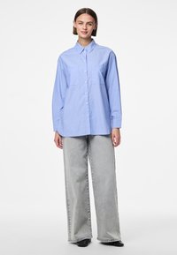 Vaaleansininen nappipaita, jossa on pystysuoria valkoisia raitoja, pitkät hihat ja kaulus, yhdistettynä harmaisiin leveälahkeisiin farkkukankaisiin housuihin.