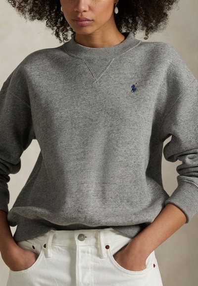 Sieviete, tērpusies pelēkā sporta džemperī ar nelielu zilu izšūtu logo, rokām kabatās baltās biksēs, stāvot pret neitrālu fonu.