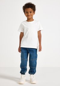 Χαμογελαστό νεαρό αγόρι με σγουρά μαλλιά, που φοράει λευκό T-shirt, μπλε τζιν παντελόνι φόρμας και λευκά αθλητικά παπούτσια, στέκεται μπροστά σε απλό φόντο.