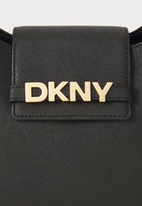 Juodas rankinukas pagamintas iš tekstūruotos sintetinės medžiagos su magnetiniu uždarymu. Priekyje yra auksinės spalvos "DKNY" logotipas.