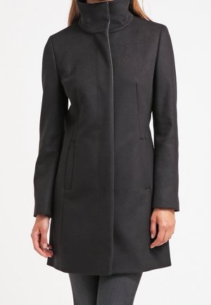 Kobieta ubrana w czarny, sięgający kolan płaszcz z wysokim kołnierzem i ukrytym zapięciem z przodu, stojąca na białym tle.