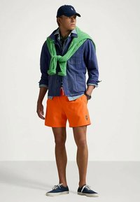 Jeansowa kurtka na jasnoniebieskiej koszuli, zielony sweter związany na szyi, pomarańczowe szorty i niebieskie płócienne buty. Swobodny strój z kontrastującymi kolorami.