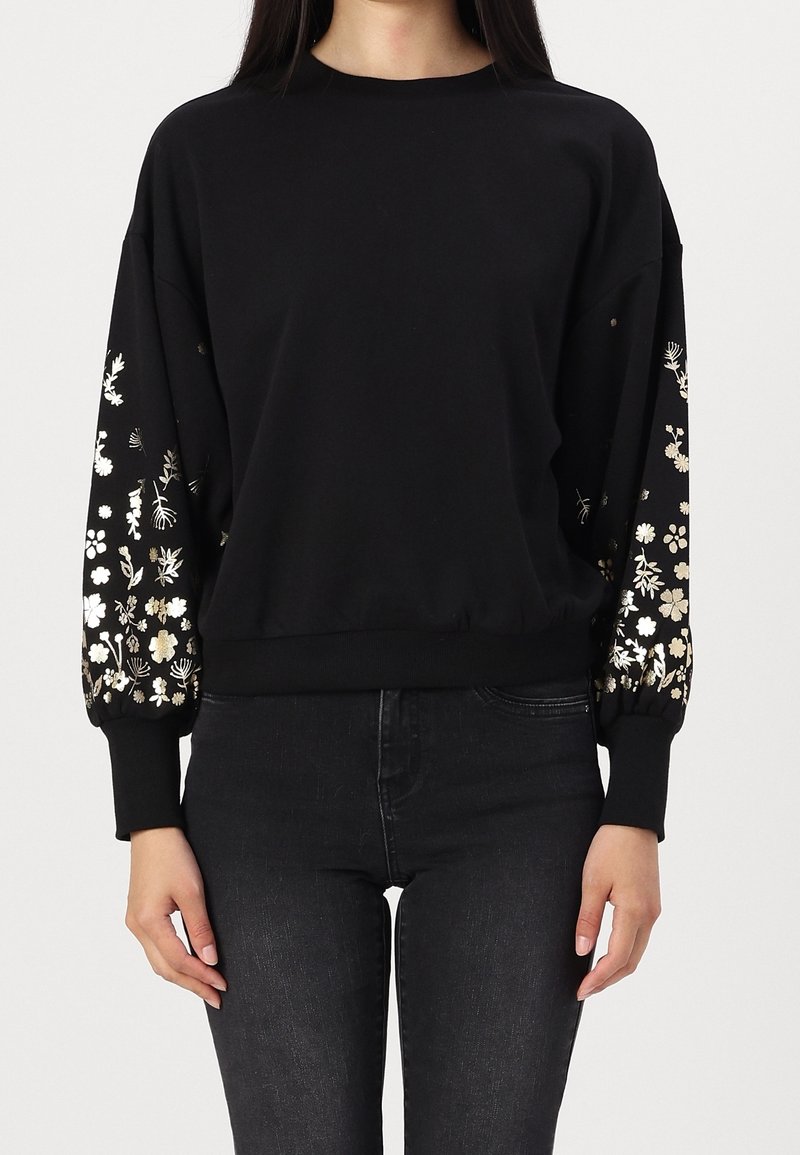 Femme portant un haut noir à manches longues avec des motifs floraux dorés et blancs sur les manches, associée à un pantalon noir.