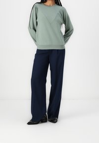 Sudadera verde con un diseño en forma de V y pliegues en los hombros, combinada con pantalones de pierna ancha en azul marino y zapatos negros de punta.