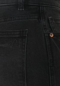 Gros plan sur un jean en denim noir montrant les poches arrière, la passoire de ceinture, les détails de couture et un rivet en cuivre sur un coin de poche.