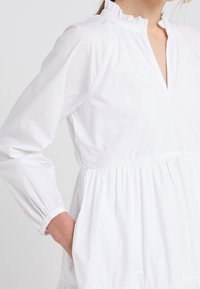 Bílá bavlněná blůza s dlouhými rukávy, nařaseným výstřihem a nabraným pasem. Obsahuje částečné zapínání a boční kapsy.