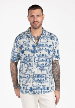 Mężczyzna z krótkimi włosami i tatuażami, ubrany w białą i niebieską krótki rękaw koszulę na guziki z wzorem oraz jasne spodnie.