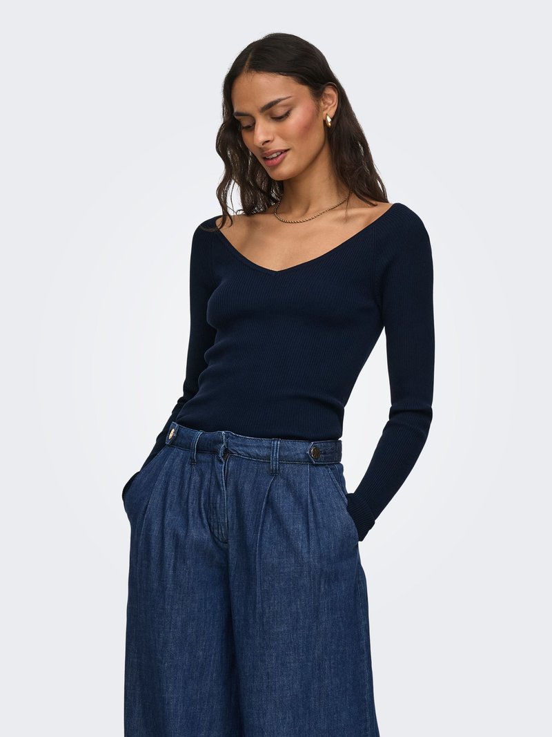 Femme portant un haut bleu marine à manches longues et col en V, ainsi qu'un pantalon en denim ample taille haute avec les mains dans les poches, devant un fond uni.