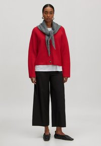 Rote Strickjacke mit weiter Silhouette, ausgestattet mit großen Knöpfen. Getragen über einem weißen Oberteil, kombiniert mit schwarzen weiten Hosen und einem grauen Schal.