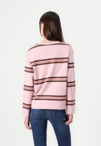 Maglione a maniche lunghe di colore rosa con strisce orizzontali marroni e rosse. Presenta un design rilassato e un ampio scollo. Abbinato a jeans in denim blu.