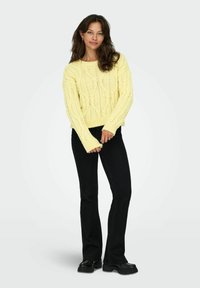 Pull en tricot jaune à motif en fausse torsade, avec manches longues, associé à un jean évasé noir et des bottes noires à semelle épaisse. Fond clair uni.