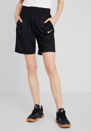 Person iført sorte Nike sportsshorts med hvidt logo, en hvid top og sorte atletiske sko med gummisåler, står med hænderne i lommerne.