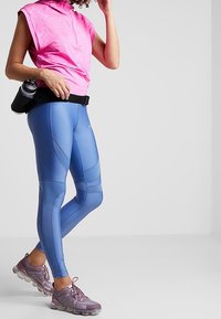 Jasnoróżowy top bez rękawów w połączeniu z niebieskimi legginsami sportowymi z fakturowanym wzorem, dopełniony fioletowymi butami sportowymi. Czarny pasek w talii z butelką.