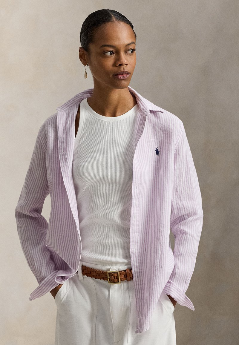 Ανοιχτό μωβ ριγέ πουκάμισο, μακριά μανίκια, ανοιχτός γιακάς, συνδυασμένο με λευκό τοπ και κρεμ παντελόνι, συμπληρωμένο με καφέ πλεκτή ζώνη.