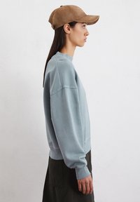  Lichtblauwe katoenen sweatshirt met lange mouwen en geribbelde manchetten, gecombineerd met een tan suède pet, zijwaarts gedragen tegen een neutrale achtergrond.
