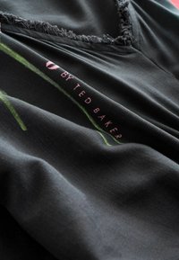 Zwarte stof met een zachte textuur, voorzien van een groen bloemenpatroon en de tekst "by Ted Baker" in het roze, nabij de onderkant.