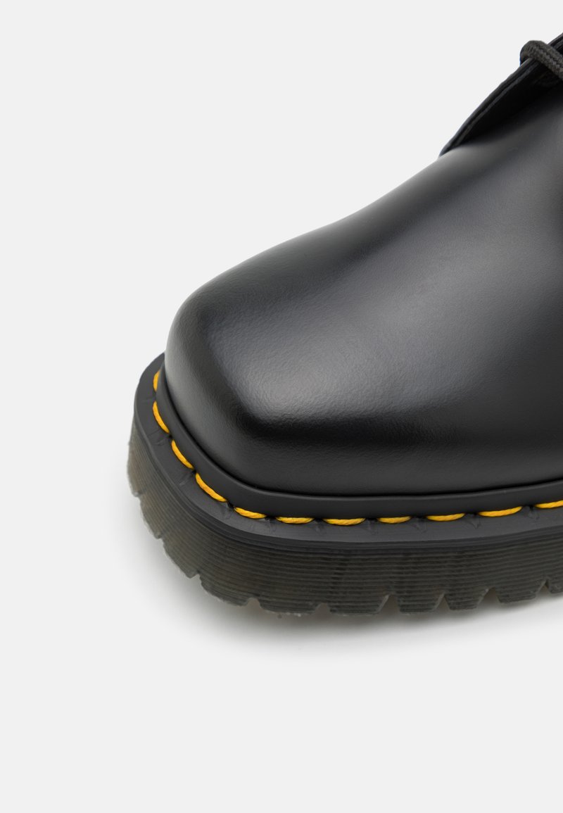 Zapato de cuero negro con punta redondeada, que destaca una suela texturizada y costuras amarillas visibles a lo largo del borde. Ensamblaje de cordones de perfil bajo.