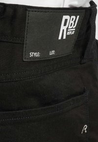 Nærbilde av baklommen på svarte denimjeans med en svart etikett som viser hvit "RBJ REPLAY"-logo og tomme felt for stil og parti.