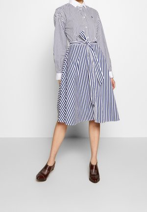 Robe chemise rayée en bleu et blanc, avec des manches longues, un col et une taille nouée. Assortie à des chaussures marron, sur un fond uni.