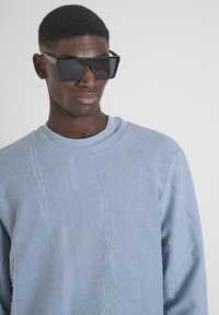 Gafas de sol negras de gran tamaño con diseño angular; combinadas con un suéter texturizado azul claro que presenta detalles deshilachados y cuello redondo.