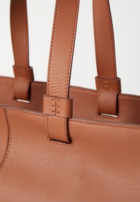 Sac à main en cuir tan avec deux poignées robustes, présentant une texture lisse et des coutures détaillées au niveau des attaches des poignées.
