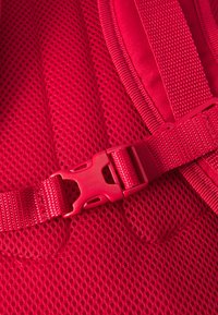 Red mesh fabric with a textured finish, featuring a plastic buckle and adjustable strap. The design emphasizes breathability and support.