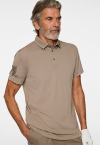 Mężczyzna ubrany w beżową koszulkę polo z czarnym wzorem w kratę na rękawie i spodnie do kompletu, stojący z jedną ręką w kieszeni i białą rękawiczką na drugiej dłoni.