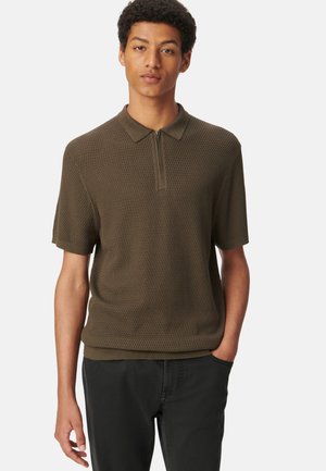 Polo de color verde olivo con un tejido texturizado de punto, mangas cortas y un diseño de cuello con media cremallera. Combinado con pantalones oscuros.