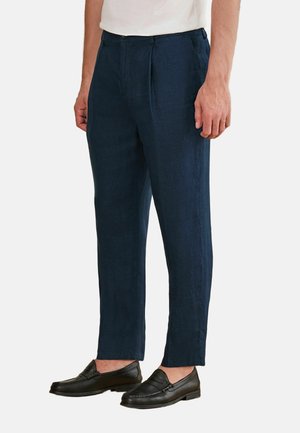 Mann trägt dunkelblaue plissierte Hose, weißes Hemd und schwarze Ledermokassins aus Leder, steht vor einem schlichten weißen Hintergrund.