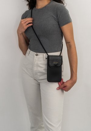 Persoon draagt een grijs shirt met korte mouwen en witte broek, houdt een zwart crossbody telefoontasje vast met een getextureerde klep en een gouden logo.