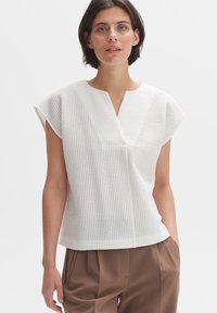 Witte textuur top met korte cap mouwen, een V-halslijn en een losse pasvorm, gecombineerd met lichtbruine geplooide broeken.