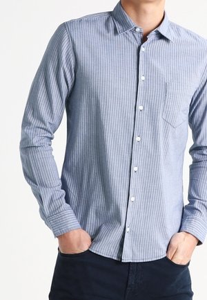 Homme portant une chemise à rayures verticales bleu clair à manches longues avec une poche poitrine, mains dans les poches d’un pantalon foncé.