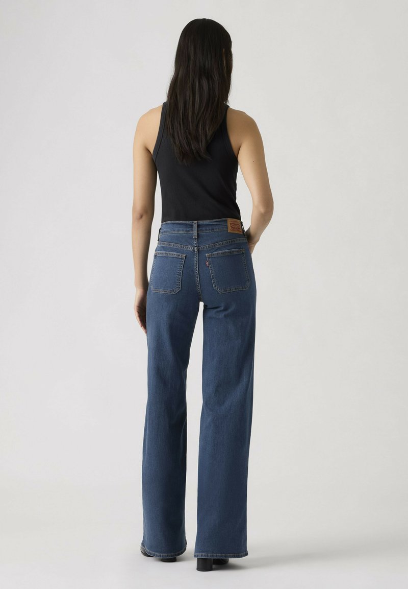 Vrouw met lang donker haar, gekleed in een zwart mouwloos topje en een hoge taille, rechte blauwe spijkerbroek, staande met haar rug naar voren gericht.