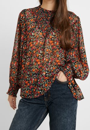 Vrouw draagt een doorschijnende zwarte blouse met oranje en roze bloemenprint en lange mouwen, gecombineerd met donkerblauwe spijkerbroek.