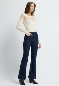 Jasna beżowa bluzka z długim rękawem w prążki, z przezroczystymi paskami; połączona z ciemnoniebieskimi spódnicami dzwonami z teksturowanym kropeczkowym wzorem i wysokim stanem.