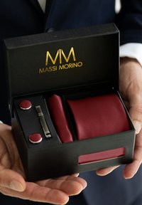 Corbata roja con un acabado texturizado, gemelos a juego y un sujetacorbatas presentados en una caja negra con letras doradas, sostenida en una mano.