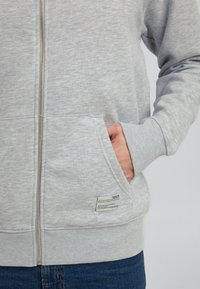 Sudadera gris con cremallera hecha de una tela suave, que cuenta con un bolsillo frontal, puños acanalados y una etiqueta cerca del dobladillo. El diseño es simple y casual.
